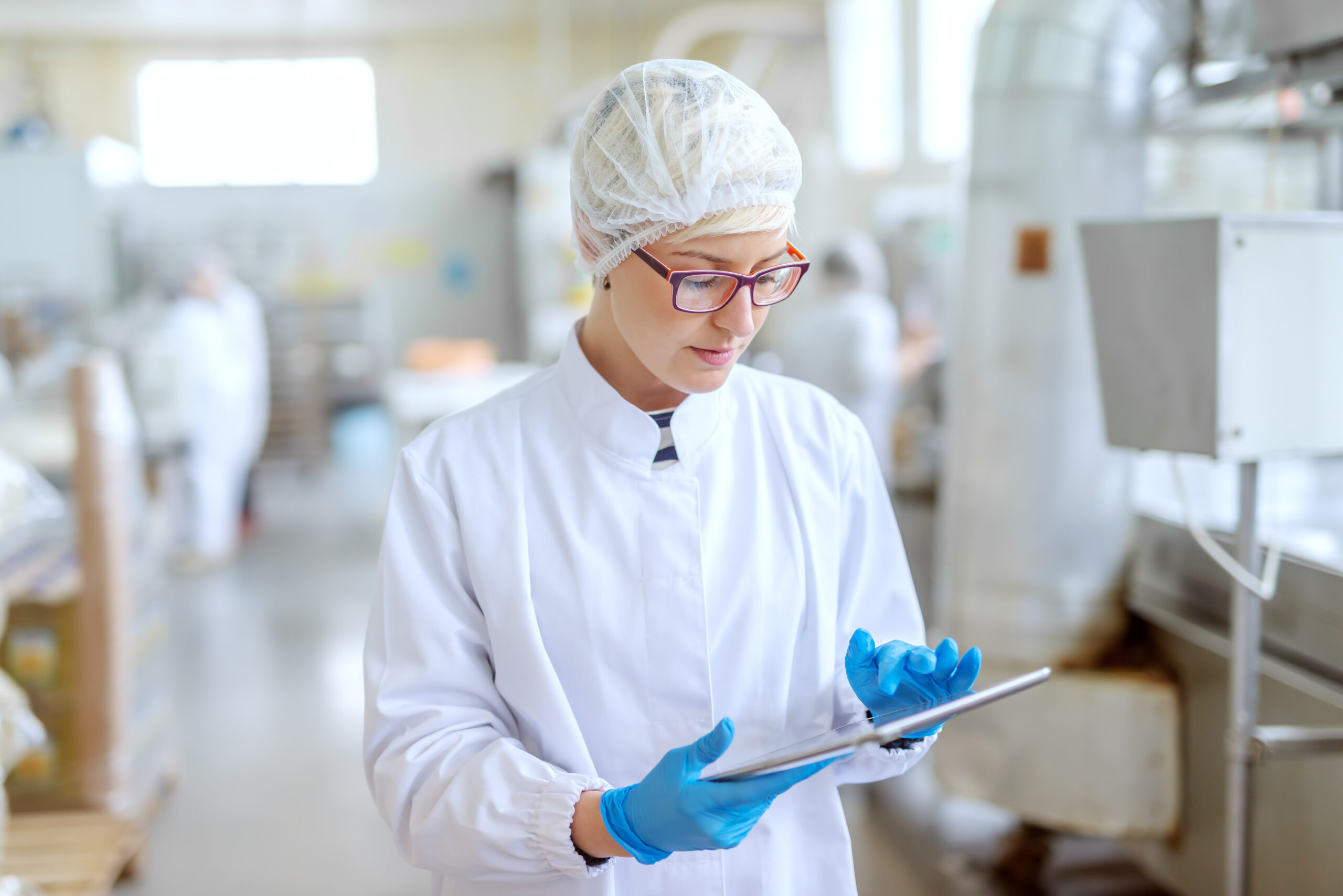 A woman in a white-coat and blue gloves in a factory looking at a device